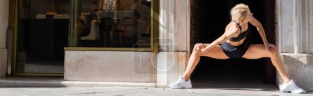 Photo pour Femme blonde en baskets blanches, haut de culture noir et short s'étirant sur la rue urbaine en Italie, bannière - image libre de droit