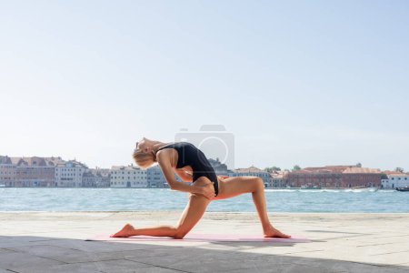 Foto de Vista lateral de la mujer estirándose mientras practica yoga en terraplén en Venecia - Imagen libre de derechos