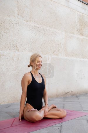 Mujer sonriente sentada en una pose de loto en una esterilla de yoga en la calle urbana de Venecia 