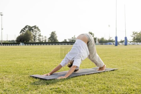 barefoot man in white t-shirt and cotton pants practicing yoga in dolphin pose on green lawn outdoors