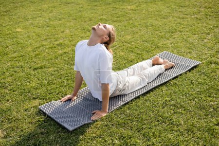 Téléchargez les photos : Homme pieds nus en t-shirt blanc et pantalon de lin pratiquant le yoga en pose de cobra sur pelouse verte - en image libre de droit