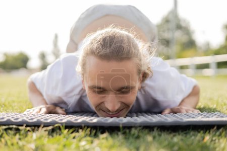 surface level of carefree man with closed eyes smiling while practicing yoga on green grass outdoors