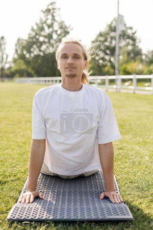 joven hombre positivo en camiseta blanca mirando a la cámara mientras practica la pose de cobra en la esterilla de yoga al aire libre