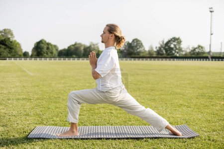 Téléchargez les photos : Vue latérale de l'homme pieds nus pratiquant le yoga en posture guerrière avec geste anjali mudra sur l'herbe verte du stade extérieur - en image libre de droit