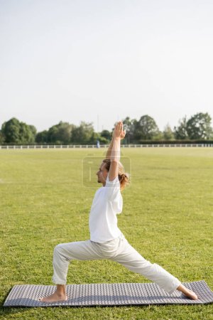 Photo for Side view of man in linen pants and white t-shirt practicing warrior pose with raised praying hands on yoga mat on green lawn - Royalty Free Image