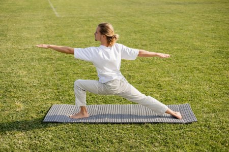 Foto de Vista lateral del joven hombre de pelo largo en camiseta blanca meditando en pose de guerrero con las manos extendidas en la esterilla de yoga al aire libre - Imagen libre de derechos