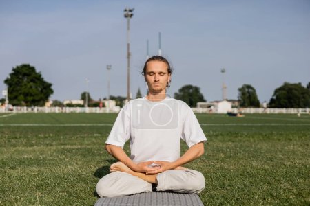 joven en camiseta blanca meditando en pose de loto con los ojos cerrados en el campo verde del estadio al aire libre