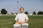 young man in white t-shirt meditating in lotus pose with closed eyes on green field of outdoor stadium puzzle #648518706