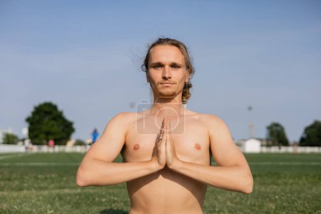 sportive shirtless man looking at camera and meditating with anjali mudra gesture outdoors