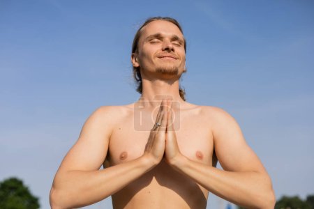 Blick auf einen unbeschwerten Mann ohne Hemd, der mit geschlossenen Augen und Anjali-Mudra-Geste vor blauem wolkenlosem Himmel meditiert