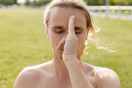 junger Yoga-Mann mit nackten Schultern und geschlossenen Augen, der die Hand vor dem Gesicht hält und draußen Pranayama-Übungen macht
