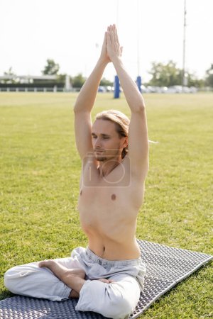 young shirtless man sitting in lotus pose with raised praying hands while practicing yoga outdoors