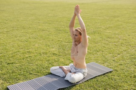 happy shirtless man in cotton pants sitting in lotus pose with raised hands while meditating on yoga mat and green grass outdoors