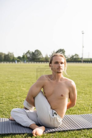 barefoot shirtless man in linen pants looking away while practicing yoga in sage pose on green field of outdoor stadium