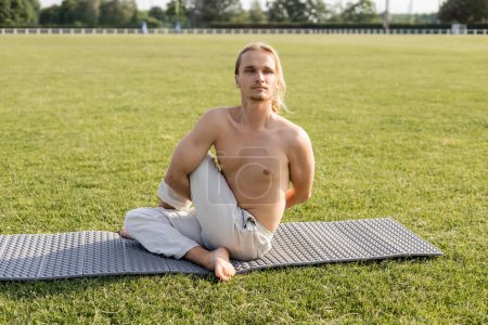 Hemdloser Mann in Baumwollhose sitzt in Salbeipose und schaut weg, während er Yoga auf grünem Rasen im Freien praktiziert