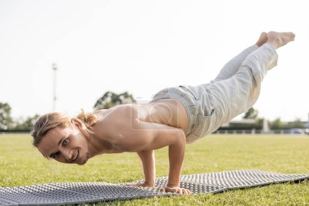 glückliches Hemd in Baumwollhose Mann schaut in die Kamera, während er Yoga in Pfau-Pose auf Rasen-Stadion praktiziert