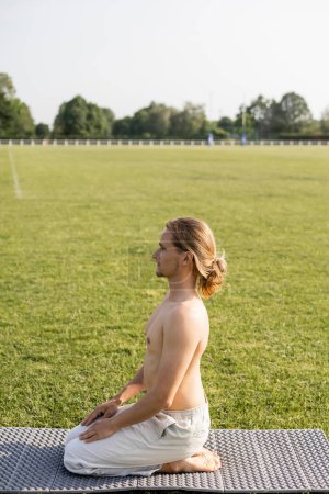 Foto de Vista lateral del hombre descalzo sin camisa sentado en la pose del rayo mientras medita en el campo verde en la esterilla de yoga - Imagen libre de derechos