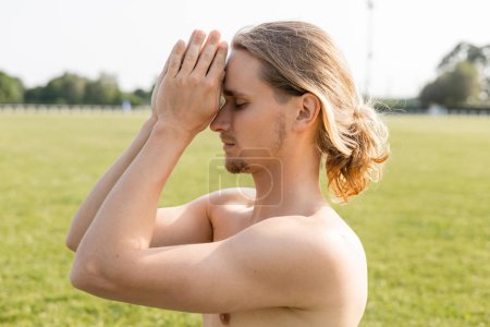 Téléchargez les photos : Vue latérale de l'homme aux cheveux longs torse nu méditant avec des mains priantes près de la tête à l'extérieur - en image libre de droit
