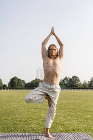 shirtless long haired man in linen pants meditating in tree pose with raised praying hands on yoga mat outdoors