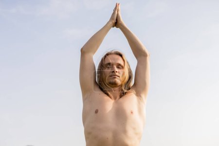 Téléchargez les photos : Vue à angle bas de l'homme torse nu avec les cheveux longs et les yeux fermés méditant avec les mains levées priant contre le ciel bleu - en image libre de droit