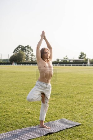 shirtless barefoot man in linen pants meditating in tree pose with raised hands on yoga mat outdoors