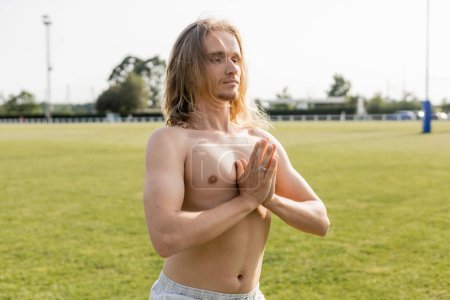 hemdlose langhaarige Yoga-Mann meditiert mit betenden Händen auf Rasen Stadion