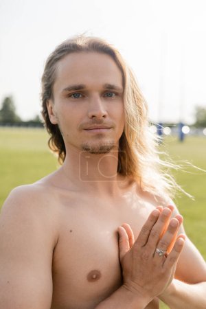 positive shirtless man with long hair meditating with anjali mudra gesture and smiling at camera outdoors