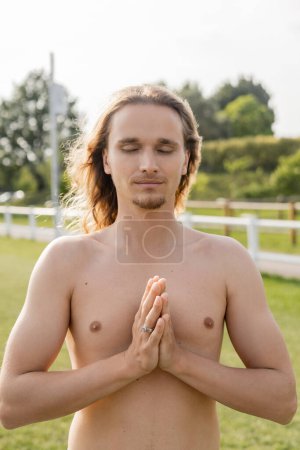 shirtless man with long hair and closed eyes showing anjali mudra gesture while meditating outdoors