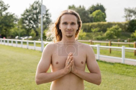 long haired and shirtless man smiling at camera while meditating with anjali mudra gesture outdoors