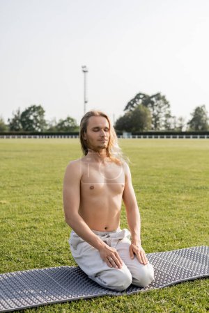 positive shirtless man in linen pants meditating in thunderbolt pose with closed eyes on green field of outdoor stadium