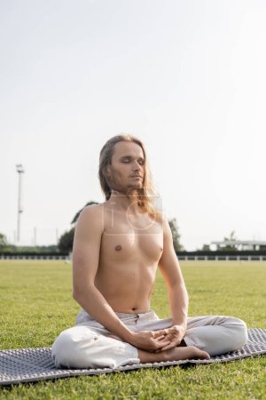 Hemdloser und sportlicher Mann mit geschlossenen Augen meditiert in lockerer Yoga-Pose auf grünem Rasen