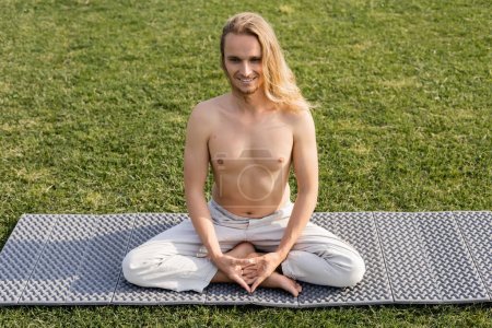 hombre sin preocupaciones sin camisa sentado en pose fácil mientras practica yoga en el campo de hierba verde