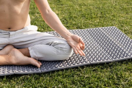 partial view of shirtless man in linen pants meditating in easy pose with gyan mudra gesture on yoga mat on green grass