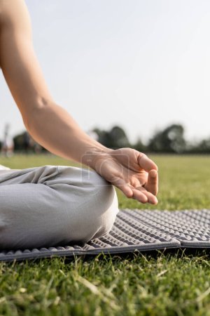 Téléchargez les photos : Vue recadrée du jeune homme méditant et montrant un geste de mudra gyan assis sur un tapis de yoga sur une pelouse verte - en image libre de droit