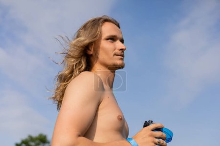 vista de ángulo bajo de hombre joven con torso sin camisa y pelo largo sosteniendo botella deportiva y mirando al aire libre
