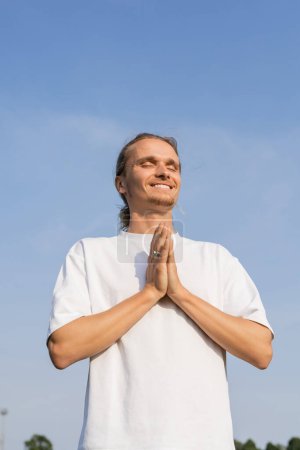 vista de ángulo bajo del hombre alegre en camiseta blanca meditando con los ojos cerrados y las manos rezando bajo el cielo despejado