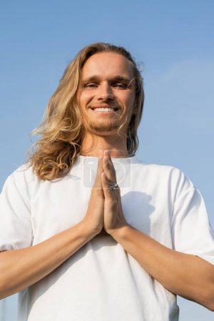vista de ángulo bajo de sonriente hombre de yoga de pelo largo meditando con las manos orantes y mirando a la cámara contra el cielo azul