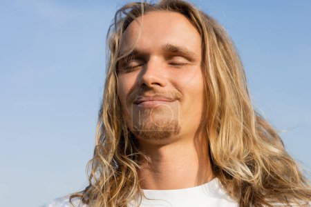 Porträt eines jungen langhaarigen Mannes, der mit geschlossenen Augen im Freien lächelt und meditiert