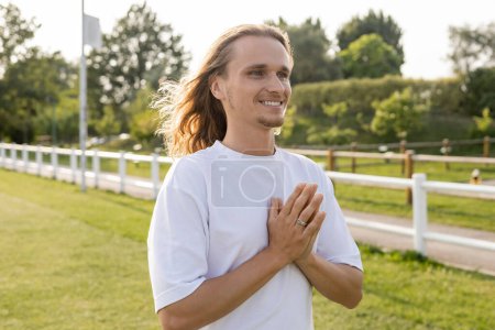 alegre hombre de pelo largo en camiseta blanca mirando hacia otro lado mientras medita con anjali mudra al aire libre