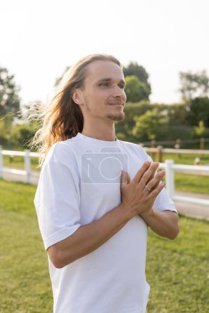 calm and carefree man with long hair looking away while meditating with anjali mudra outdoors magic mug #648519590
