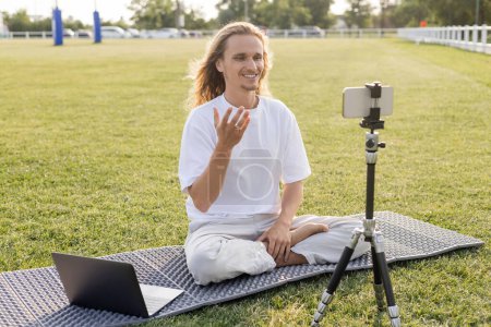 happy yoga coach sitting in easy pose near laptop and mobile phone on tripod on green lawn of outdoor stadium
