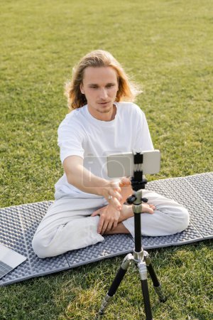 joven vlogger de yoga ajustando trípode con teléfono móvil mientras está sentado en pose fácil en campo herboso