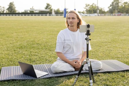 Téléchargez les photos : Professeur de yoga aux cheveux longs regardant loin tout en étant assis près de l'ordinateur portable et trépied avec téléphone portable sur le terrain du stade extérieur - en image libre de droit