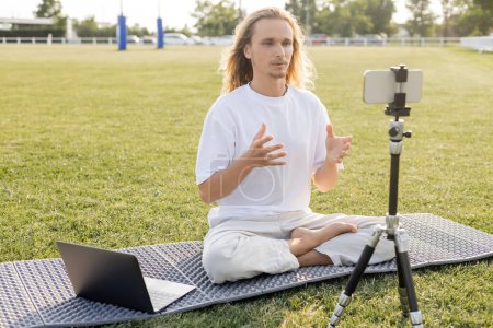 Foto de Entrenador de yoga hablando cerca de trípode con teléfono inteligente y portátil mientras está sentado en pose fácil en el estadio cubierto de hierba - Imagen libre de derechos