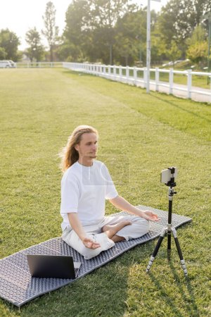 Téléchargez les photos : Professeur de yoga montrant pose facile et gestes gyan mudra assis sur tapis de yoga près du smartphone sur trépied à l'extérieur - en image libre de droit