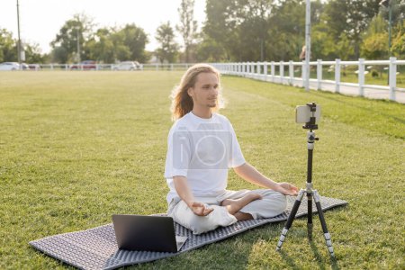 yoga vlogger meditating in easy pose near mobile phone on tripod on green lawn of outdoor stadium