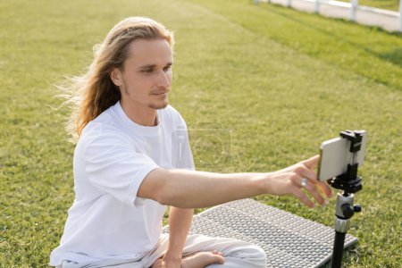 young long haired man using mobile phone while sitting on yoga mat on green grassy field 