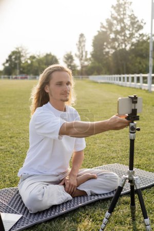yoga vlogger sitting in easy pose and adjusting tripod with smartphone while sitting on green grass outdoors