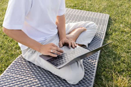 Téléchargez les photos : Vue recadrée de l'homme en utilisant un ordinateur portable tout en étant assis en pose facile sur tapis de yoga sur herbe verte - en image libre de droit