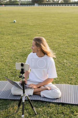 Téléchargez les photos : Coach de yoga assis avec ordinateur portable près du téléphone portable sur trépied et regardant loin sur le stade herbeux - en image libre de droit